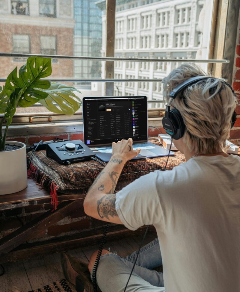 Music producer with headphones creating tracks on a laptop in a bright, indoor setting.